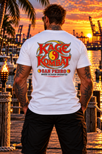 A man stands on a dock at sunset wearing a black T-shirt with a large 'Kage Kombat San Pedro' graphic on the back, with a tropical harbor and port cranes in the background.