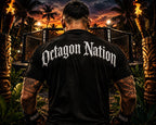 A man viewed from behind wearing a black t-shirt with 'Octagon Nation' printed in white gothic lettering across the back, standing in a tropical ring arena with tiki torches and a cage in the background.
